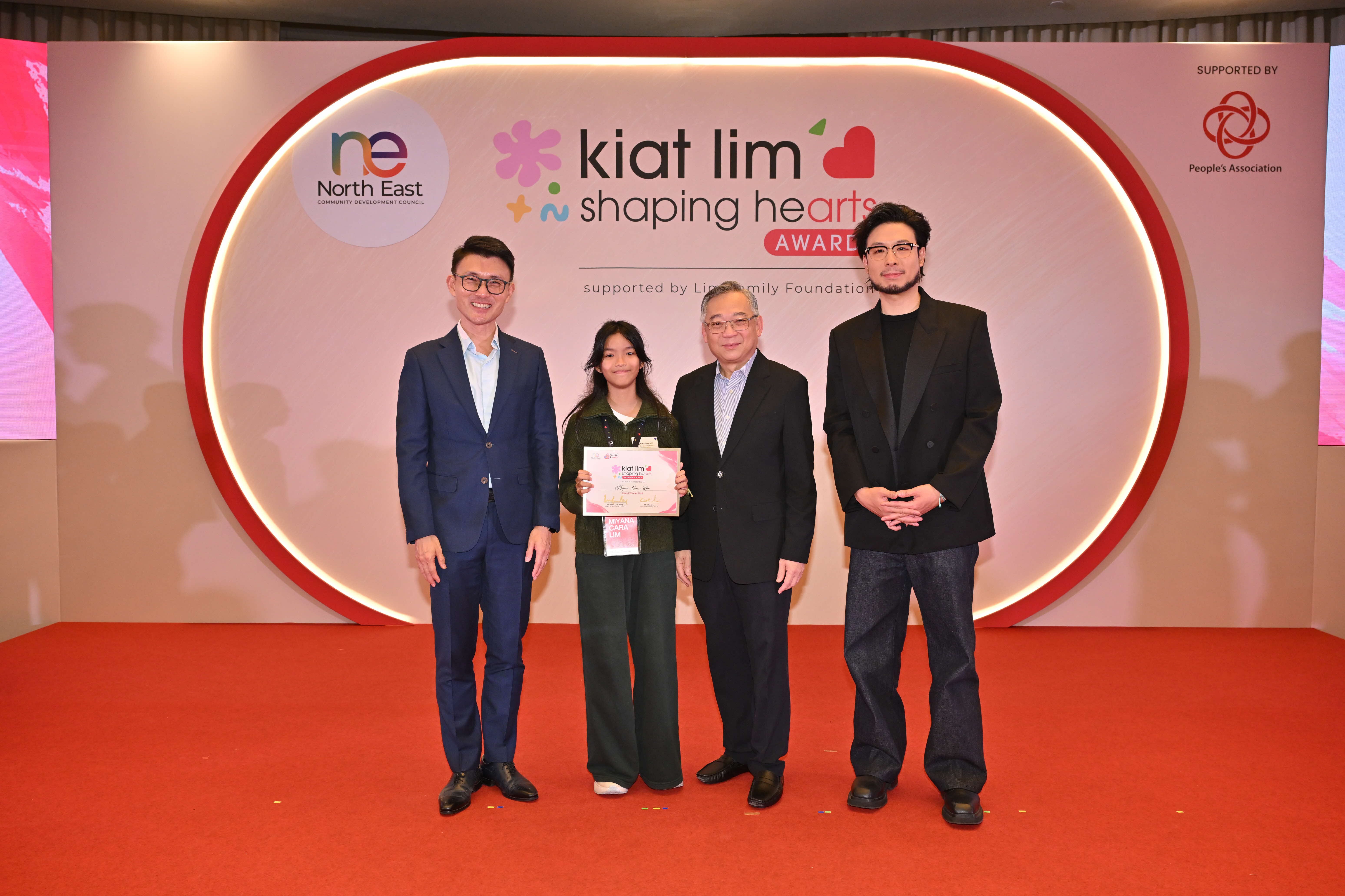 Award recipient standing on stage with the presenter, holding their certificate and posing for a group photo during the award ceremony, with a backdrop reading ‘Kiat Lim-Shaping Hearts Awards'.